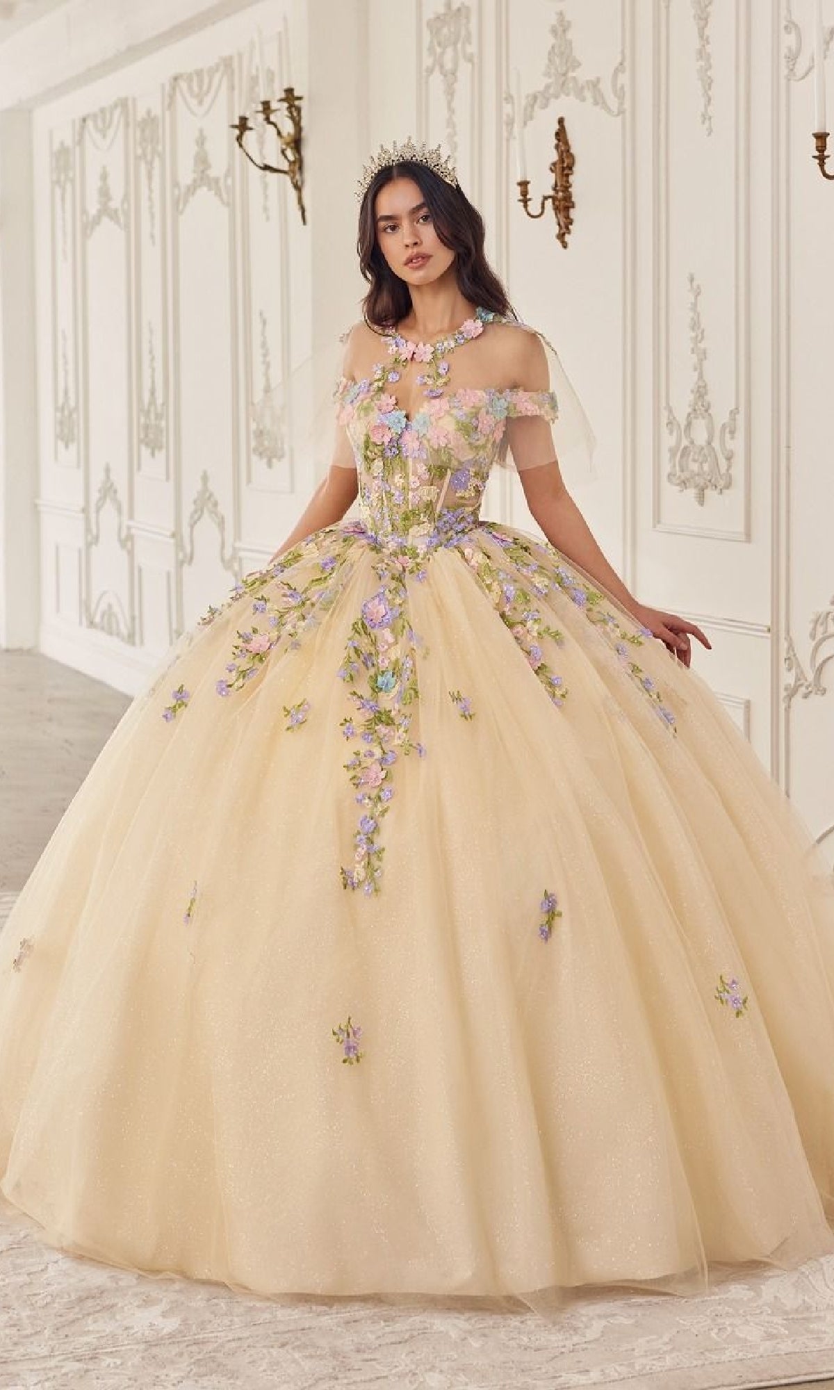 In an ornate room, a woman wears the Quinceanera Dress 15724 by Ladivine—a stunning off-the-shoulder light yellow ball gown with purple and pink floral appliqués and a tiara.